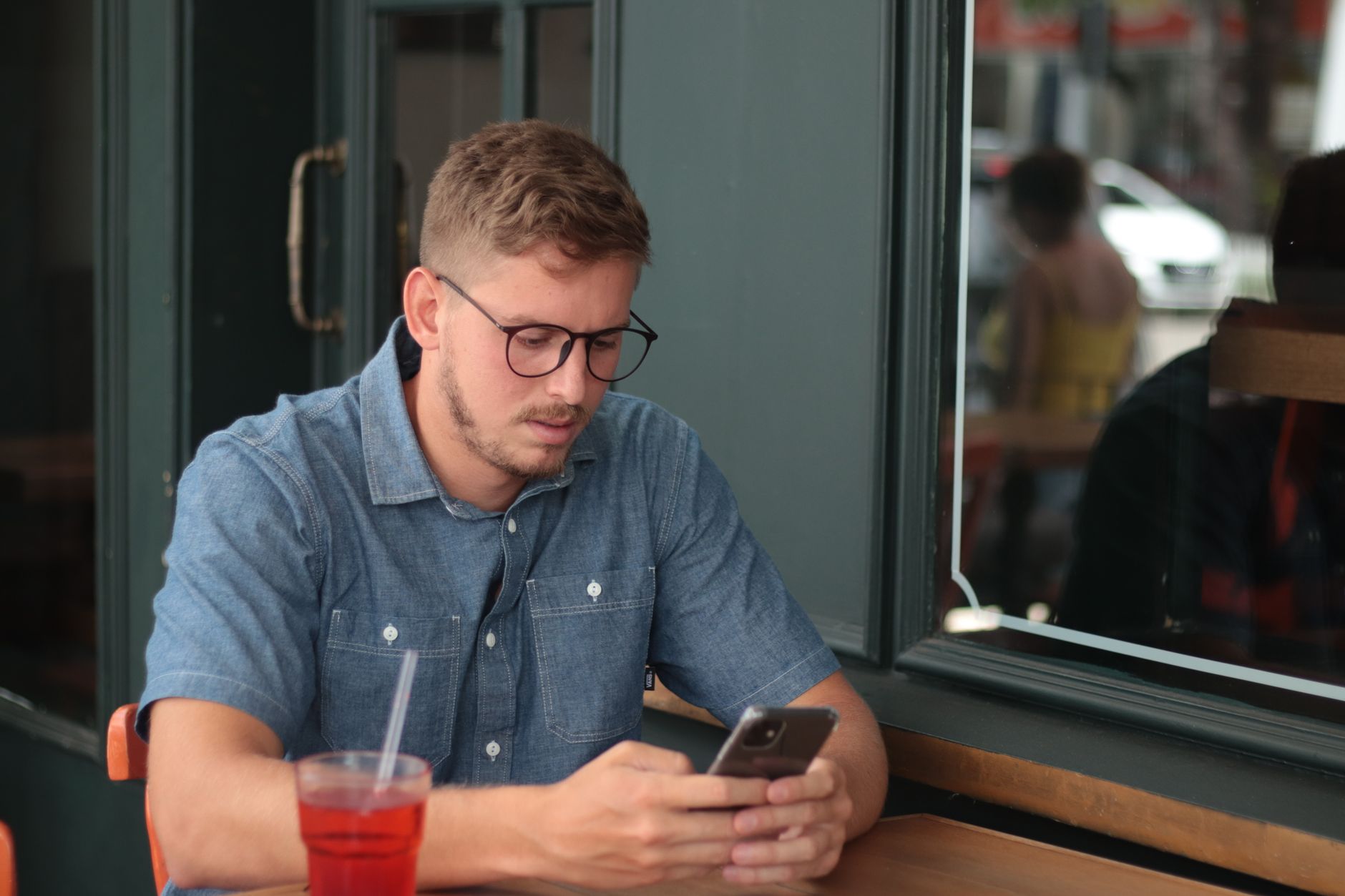 Un giovane con gli occhiali legge lo smartphone seduto a un tavolino di un caffè all'aperto
